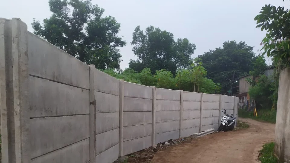 Deretan panel dan tiang pagar beton pracetak berdiri rapi di tepi jalan tanah